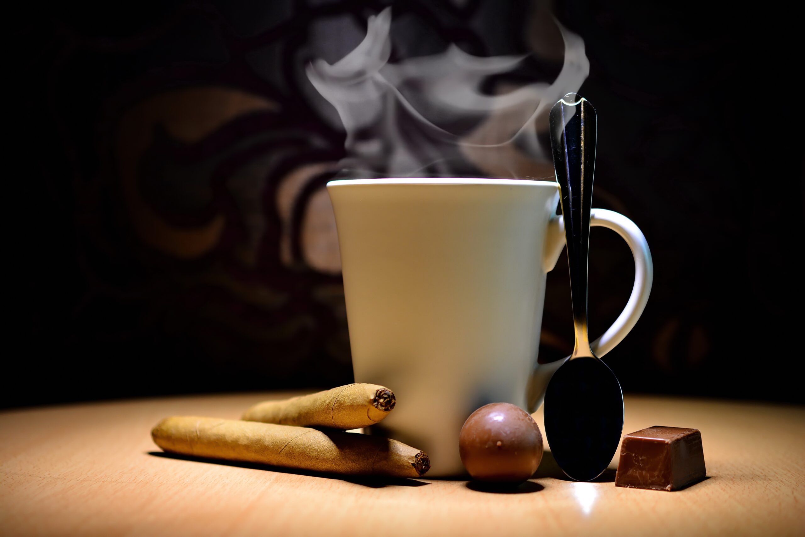 A steaming cup of coffee sits on a wooden surface, accompanied by a metal spoon, a piece of chocolate, and two cigars. The dark background highlights the warm, cozy ambiance.